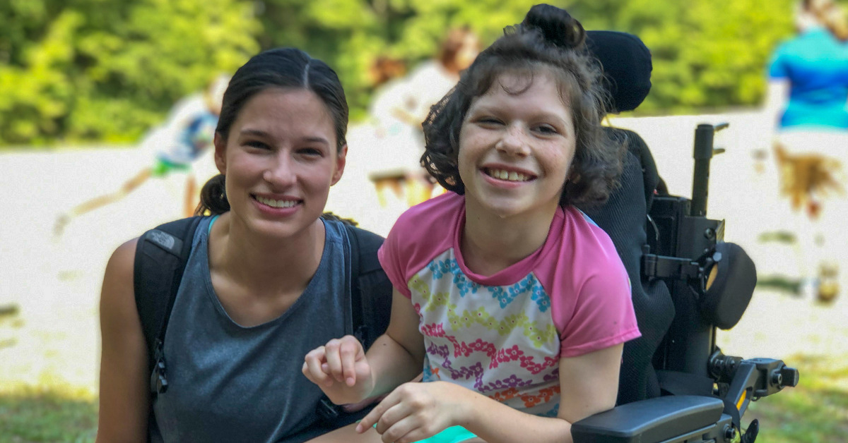A caucasian teenage girl wearing a gray tank top and who has brown hair in a pony tail is kneeling next to a female camper with short brown hair who is wearing a pink swim shirt and is in a motorized wheelchair. Both girls are smiling at the camera.