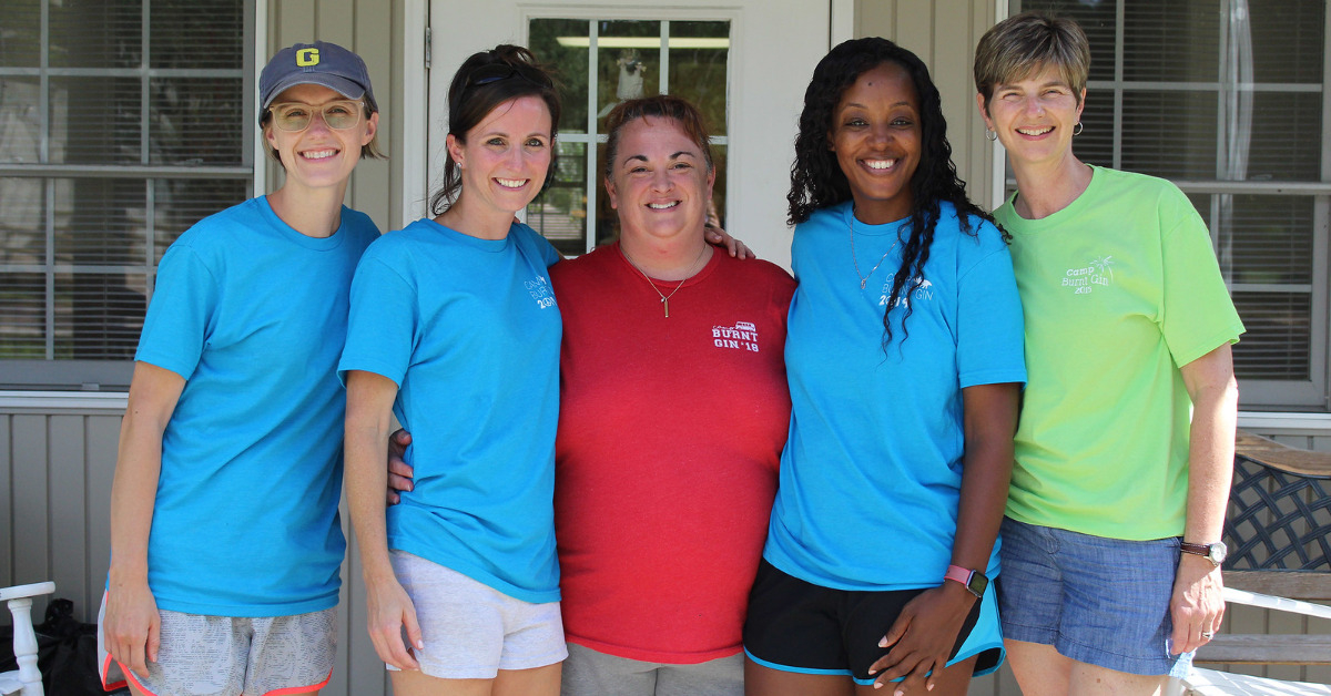 Five female staff members stand on a porch smiling at the camera. They have on Camp Burnt Gin '19 shirts and running shorts on.