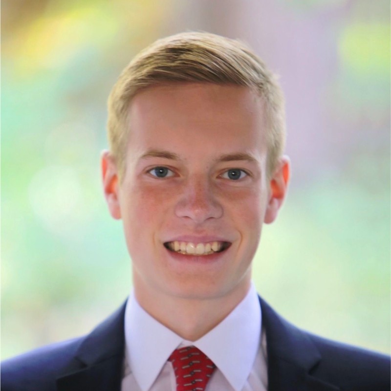 A caucasian boy with blonde hair and blue eyes is wearing a blue suit, white shirt, and red tie. The boy looks to be in his upper teens or low twenties