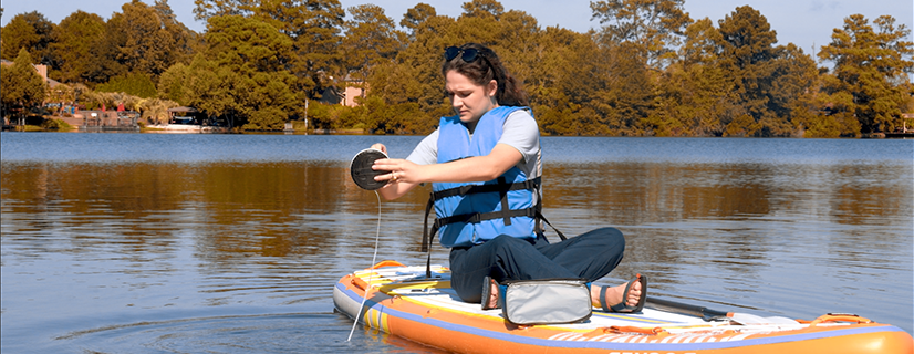 Female wearing a blue life jacket is sitting cross-legged on a raft collecting data from the lake