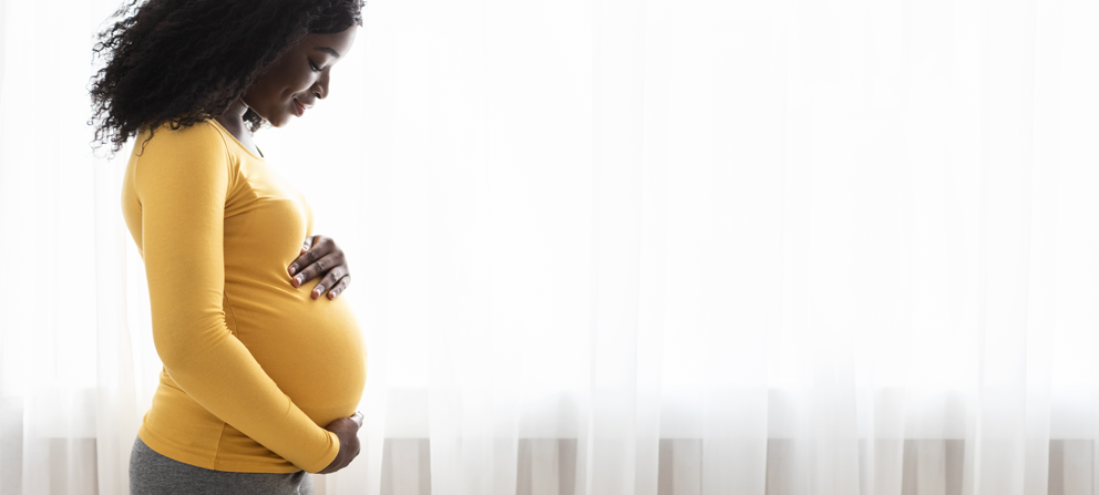 African American pregnant woman with shoulder length hair wearing a yellow long sleeve shirt and gray leggings is holding her baby bump facing to the right. She looks to be in baby's room with gray curtains and lots of natural light behind her