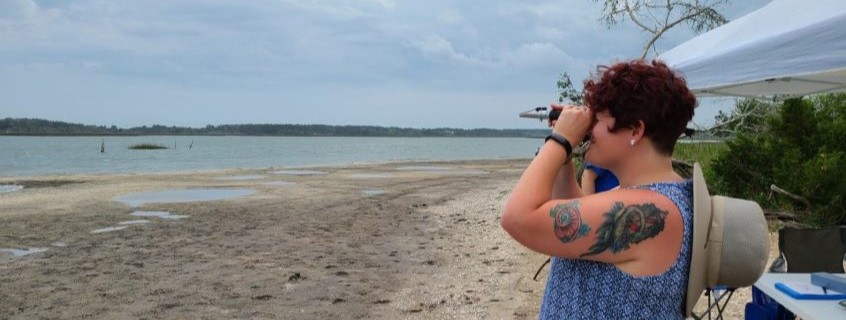 Woman with hat hanging behind her neck uses binoculars to look out into the ocean